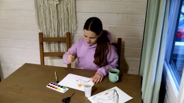 Bir kız ahşap bir masada oturur, kapalı bir ortamda suluboya boyayla resim yapar. Sanat malzemeleriyle çevrili, yaratıcılık ve konsantrasyonla dolu huzurlu bir öğleden sonranın tadını çıkarıyor..