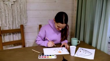 Genç bir kadın, rahat bir yerde, ahşap bir masada otururken suluboya kullanarak güzel bir sanat eseri yaratıyor..