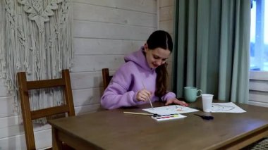 Bir kız ahşap bir masada oturuyor, suluboya resimlerle meşgul, bir fincandan yudumlarken. Doğal ışık odayı doldurur ve sıcak bir atmosfer yaratır..