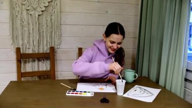 Genç bir kız suluboya resimlerle mutlu bir şekilde yaratıcı sürecini rahat bir odada, ahşap bir masada sürdürüyor. Rahat bir atmosferde oturuyor, bir bardaktan yudumluyor..