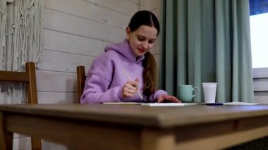 Genç bir sanatçı, sıcacık bir ahşap odanın içindeki suluboya resmine odaklanmış. Bir masada oturuyor, sıcak içkisinin tadını çıkarırken fırça kullanıyor, yaratıcı sürecine dalmış..