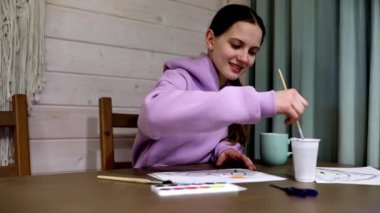 Rahat kapüşonlu bir kız, sanatsal eserlerine odaklanmış bir masada neşeyle resim yapıyor. Bir bardak yanında, fırça ve suluboya palet kullanıyor..