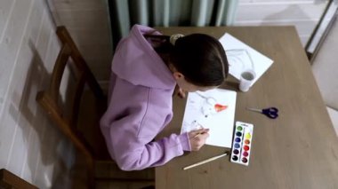 Genç bir kadın, rahat bir yerde, ahşap bir masada oturuyor, suluboya ile dikkatlice resim yapıyor. Etrafı materyallerle ve sıcak bir atmosferle çevriliyken sanatına odaklanıyor..