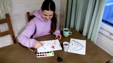 Sanat malzemeleriyle çevrili, ahşap bir masada resim yapan genç bir sanatçı, sıcak bir ortamda sanatsal çalışmalarına odaklanırken sıcak bir içkinin tadını çıkarıyor..