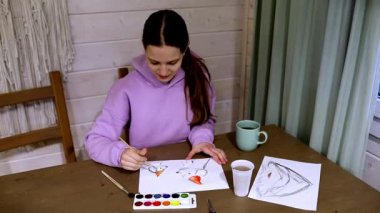 Genç bir kadın ahşap bir masada oturmuş, odaklanmış bir şekilde suluboya resim yapıyor. Yaratıcılığını yansıtırken yanında sıcak bir fincan çay içiyor..