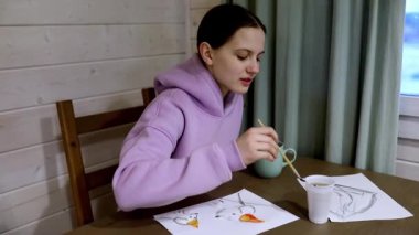 Mor kapüşonlu bir kız ahşap bir masadaki sanat eserlerine odaklanır. Çeşitli konuların çizimlerine ayrıntıları eklemek için fırça kullanıyor, sıcak bir içecekten zevk alıyor..