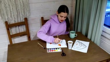 Bir insan ev içi resim yapma seansının keyfini çıkarıyor. Suluboya kullanarak, rahat bir kulübedeki ahşap bir masada güzel çizimler yapıyorlar. Yakınlarda sıcak bir içecek var, dingin atmosfere katkı sağlıyor..