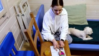 Kapüşonlu bir kız rahat bir odadaki masada renkli figürler toplamaya odaklanıyor. Yakındaki bir kanepede rahatlayan küçük bir köpek atmosfere sıcaklık katar..