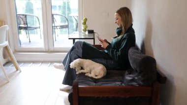 Genç bir kadın şık bir oturma odasında, akıllı telefonuna odaklanmış bir kanepede rahatça oturuyor. Küçük bir köpek onun yanında huzur içinde uzayın huzurlu atmosferinin tadını çıkarıyor..