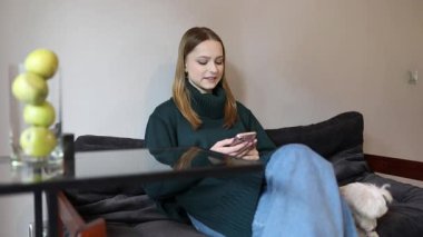 Genç bir kadın rahat bir şekilde oturma odasındaki kanepede oturuyor. Köpeği yakınlarda dinlenirken telefonuna kayıyor ve rahat bir atmosfer yaratıyor..