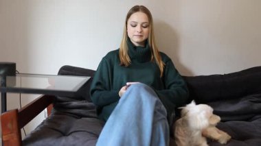 Genç bir kadın rahat bir şekilde kanepede oturur ve sessiz bir öğleden sonranın tadını çıkarır. Küçük köpeği yakınlarda, rahat oturma odasının rahat atmosferine sıcaklık katıyor..