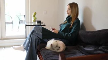 Bir kadın evinde, yanında köpeği olan bir kanepede rahatça oturmaktan zevk alır. Oturma odası aydınlık, dekoratif eşyalarla dolu küçük bir masa var..