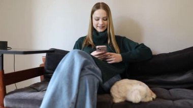 Genç bir kadın sıcak bir ortamda koltuğunda rahat bir şekilde, akıllı telefonundan süzülüyor. Yanında küçük bir evcil hayvan, rahat bir atmosfer yaratıyor..