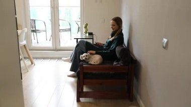 Bir kadın, küçük bir köpek kucağında dinlenirken ahşap bir banka rahatça oturur. Güneş büyük pencerelerden parlayarak sıcak bir atmosfer yaratır..