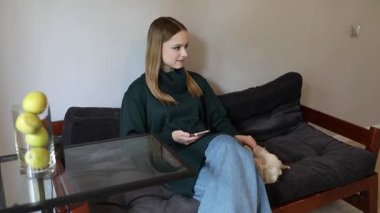 Rahat bir kazak giymiş genç bir kadın evde huzurlu bir öğleden sonranın tadını çıkarıyor. O rahat bir kanepede oturuyor, telefonuyla meşgul, yanında küçük bir köpek yavrusu dinleniyor..