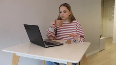 Genç bir kadın, modern bir evde bir masada oturur, bilgisayarına odaklanarak kahve içer ve kurabiye yer. Sıcak atmosfer rahat bir iş ortamı yaratıyor..