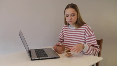 Genç bir kadın, sakin bir mekanda masada oturup laptopuna odaklanarak bir şeyler atıştırıyor ve içiyor. Rahatlık ve verimliliği harmanlayan huzurlu bir an..