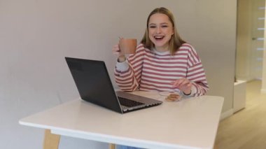 Genç bir kadın parlak bir masada oturuyor, kahvesini yudumluyor ve dizüstü bilgisayarına odaklanarak gülümsüyor. Yakınlarda bir tabak kurabiye var, sıcak ve rahat bir çalışma ortamı yaratıyor..