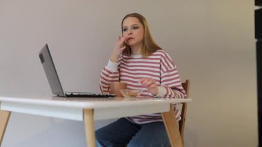 Genç bir kadın bir masada oturur ve aperatiflerin tadını çıkarırken dizüstü bilgisayarına odaklanır. Çizgili bir kazak giyiyor ve çalışma odasında rahat görünüyor..