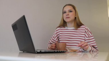 Genç bir kadın bir masada oturuyor, kahvesini yudumluyor ve laptopunu karıştırıyor. Onun yanında, küçük bir kase kurabiye bekliyor. Parlak ve basit dekor rahatlatıcı atmosfere katkı sağlar..