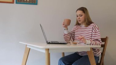 Genç bir kadın beyaz bir masada oturuyor, kahvesini yudumluyor ve laptopunu karıştırıyor. İyi ışıklandırılmış, davetkar, üretkenlik için mükemmel bir yerde odaklanmış ve rahatlamış görünüyor..