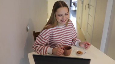 Genç bir kadın sıcak bir mutfakta beyaz bir masada oturur, kahvesini yudumlar ve kurabiyelerin tadını çıkarır. Dizüstü bilgisayarına odaklanıyor, sessiz bir anda kendini işine veriyor..