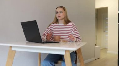 Bir kadın modern bir iç mekanda bir masada oturur, kahvesini yudumlar ve laptopuyla işe koyulur. Atıştırmalık yerken rahatlamış ve odaklanmış görünüyor..