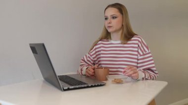 Genç bir kadın rahat bir ortamda beyaz bir masada oturmuş laptopuna odaklanarak sıcak bir içecek yudumluyor. Yanında atıştırmalık bir tabak var, verimli bir sabah olduğunu gösteriyor..