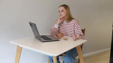 Genç bir kadın bir masada oturuyor, bir fincandan kahve yudumluyor ve özenle laptopuyla çalışıyor. Çizgili bir bluz giyiyor ve sakin bir ev ortamı ile çevrili vazifelerine odaklanmış görünüyor..