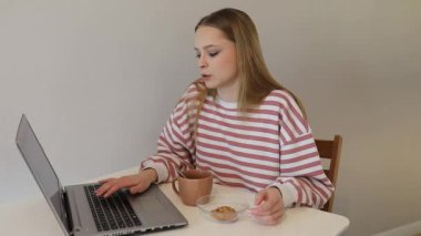 Genç bir kadın rahat bir odada bir masada oturuyor, dizüstü bilgisayarına odaklanmış. Sıcak bir içecek ve hafif bir atıştırmalık, dengeli bir iş ve rahat bir ortam içinde dinlenmeyi sever..