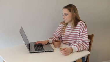 Genç bir kadın laptopuna odaklanmış bir masada oturuyor, günlük giysiler giyiyor. Sıcak bir ev ortamında yanında bir fincan ve küçük bir kase abur cubur var..