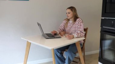 Genç bir kadın dizüstü bilgisayarına odaklanmış, parlak bir mutfak masasında oturuyor. Sıcak bir fincan kahvenin keyfini çıkararak evinde rahat ve üretken bir atmosfer yaratıyor..