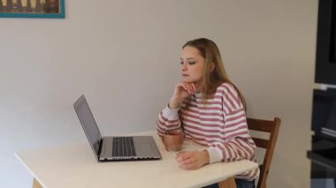 Genç bir kadın masada oturuyor, dizüstü bilgisayarına odaklanmış, rahat bir iç mekanda sıcak bir içecek yudumluyor. Basit ve davetkar bir ortamda işine dalmış gibi görünüyor..