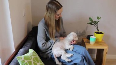 Genç bir kadın kanepede oturuyor, kucağında küçük bir köpeği nazikçe tutuyor, rahat bir anın tadını çıkarıyor bitkiler ve yumuşak dekorlarla dolu bir oturma odasında..