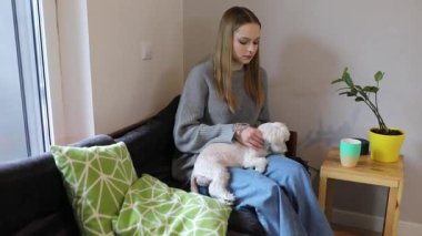 Genç bir kadın kanepede rahatça otururken küçük köpeğini nazikçe okşar. Güneş ışığı yakındaki bir pencereden süzülerek oturma odasında sıcak bir ortam yaratır..