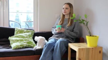 Genç bir kadın bir fincan çay içerken rahat bir kanepede dinlenir. Yanında küçük bir köpek oturuyor ve bir kapalı alan bitkisi davetkar alana biraz yeşillik katıyor..