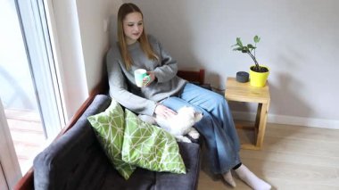 Genç bir kadın, yanında küçük bir köpekle kanepede otururken evinde çay yudumlayarak rahatlatıcı bir öğleden sonranın tadını çıkarıyor. Bir bitki rahat uzaya hayat katar..