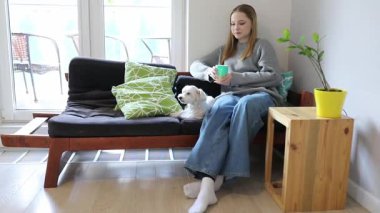 Genç bir kadın yastıklarla çevrili bir kanepede rahatça oturur, küçük köpeği yanında dinlenirken içkisini yudumlar. Huzurlu atmosfer rahatlamayı ve rahatlığı davet eder..
