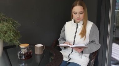 Genç bir kadın sessizce balkonda oturur, çay içerken kitap okur. Huzurlu ortamda küçük bir masa, yeşil bir bitki ve sabah güneşi yer alıyor..
