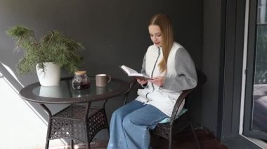 Balkonda huzurlu bir sabah geçiren bir kadın kahvesini yudumlarken kitap okuyor. Doğal ışık, bitkiler ve mobilyaların yarattığı rahat atmosferi vurguluyor.