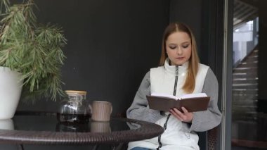 Genç bir kadın teras masasında oturuyor, sıcak bir fincan kahvenin tadını çıkarırken kendini bir kitaba kaptırmış. Modern çevre rahat ve davetkar bir atmosfer yaratır..