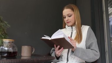 Genç bir kadın küçük bir masada rahatça oturur, kahve içerken kitap okur. Rahatlamış ve meşgul görünüyor. Dışarıdaki huzurlu atmosferin tadını çıkarıyor..