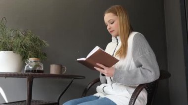 Bir kadın terasta rahatça oturup sıcak bir içecek yudumlarken kitap okumaya dalmış. Konforlu ortam yeşillik ve sakin bir atmosfer içeriyor. Rahatlamak için mükemmel..