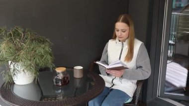 Bir kadın açık havada küçük bir masada oturuyor, okumaya dalmış bir şekilde. Etrafı yeşillik ve huzurlu bir çevreyle çevrili, rahatken sıcak bir içeceğin tadını çıkarıyor..