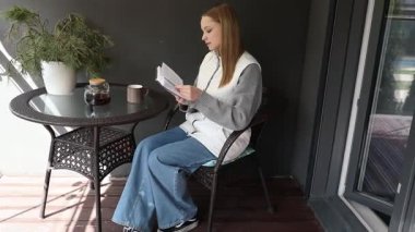 Genç bir kadın rahatça balkonunda oturuyor, kahvesini yudumlarken bir kitaba dalmış. Sakin ortam ve güneş ışığı mükemmel bir sabah atmosferi yaratır..