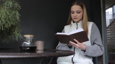 Genç bir kadın, sakin bir açık hava ortamında şık bir masada oturur, kahvesini yudumlarken bir kitaba daldırılır. Hava sakin ve öğleden sonra ışığında davetkar..