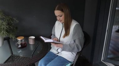 Genç bir kadın balkonda huzurlu bir şekilde oturuyor, okumaya dalmış. Çay içer ve defterine notlar yazar, sessiz bir anın tadını çıkarır..