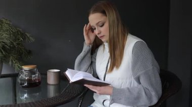 Sakin bir kafede, genç bir kadın sıcak bir içeceğin tadını çıkarırken kitabına dikkatle odaklanır. Yumuşak ışıklandırma ve rahat atmosfer rahatlamak için mükemmel bir nokta yaratır..