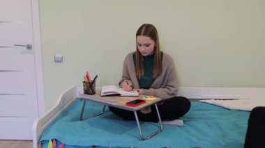 Bir öğrenci yatakta bacak bacak üstüne atarak oturur ve bir deftere yazmaya odaklanır. Renkli kırtasiye malzemeleri ve akıllı telefon yakınlarda, rahat bir çalışma ortamı yaratıyor..