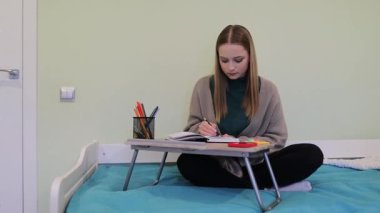 Sıcak bir yatak odasında, genç bir kadın yatağında bacak bacak üstüne atarak oturur ve derslerine odaklanır. Etrafı renkli kırtasiyelerle çevriliyken defterine notlar alır..